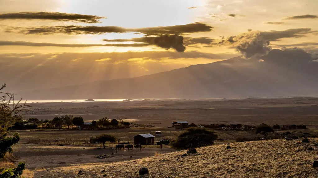 Sun rays break through clouds over a rural landscape with scattered trees, cattle, and small houses—perfect scenery for a first-time safari—with mountains and a body of water in the background.