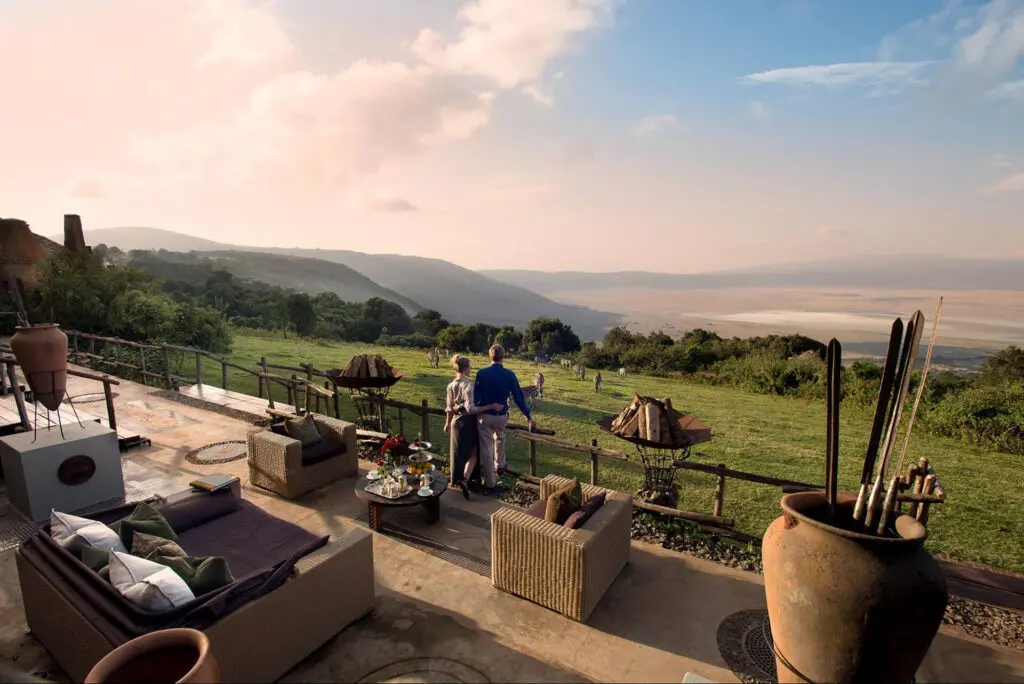 Two people stand at a fenced terrace overlooking a vast valley and distant hills, evaluating and comparing Tanzania tour packages, with outdoor furniture and decorative items in the foreground.