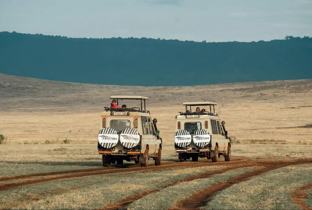 Two safari vehicles with zebra-patterned covers drive on a dirt road across a grassy plain. Hills rise in the background beneath a partly cloudy sky—an inviting scene for those looking to compare safari packages or explore Tanzania tour packages.