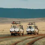 Two safari vehicles with zebra-patterned covers drive on a dirt road across a grassy plain. Hills rise in the background beneath a partly cloudy sky—an inviting scene for those looking to compare safari packages or explore Tanzania tour packages.
