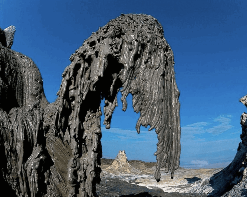 Thick, gray mud flows and hardens into unusual shapes at a mud volcano site in Tanzania under a clear blue sky, with a distant rocky outcrop—an incredible spot to visit for adventurous travel enthusiasts.