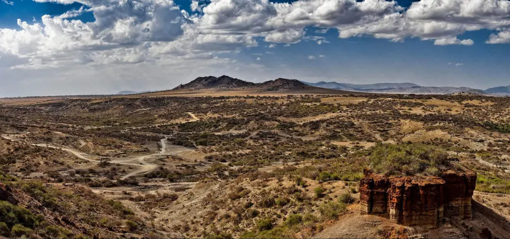 Arid landscape with sparse vegetation, dirt roads, rocky hills, and a partly cloudy sky, featuring a prominent cliff in the foreground—an ideal scene for travel enthusiasts looking to visit Tanzania’s rugged beauty.