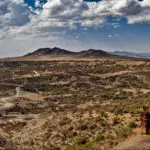 Arid landscape with sparse vegetation, dirt roads, rocky hills, and a partly cloudy sky, featuring a prominent cliff in the foreground—an ideal scene for travel enthusiasts looking to visit Tanzania’s rugged beauty.