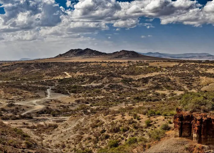 Arid landscape with sparse vegetation, dirt roads, rocky hills, and a partly cloudy sky, featuring a prominent cliff in the foreground—an ideal scene for travel enthusiasts looking to visit Tanzania’s rugged beauty.