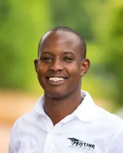 Smiling man in a white "Pristine Adventures and Safaris" shirt stands outdoors, representing Adventure Travel, with a blurred green background.