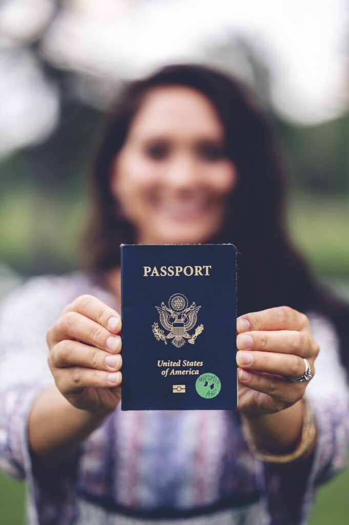 A person holds out a United States passport towards the camera, with the cover and seal clearly visible—perhaps preparing to apply for a Tourist Visa.