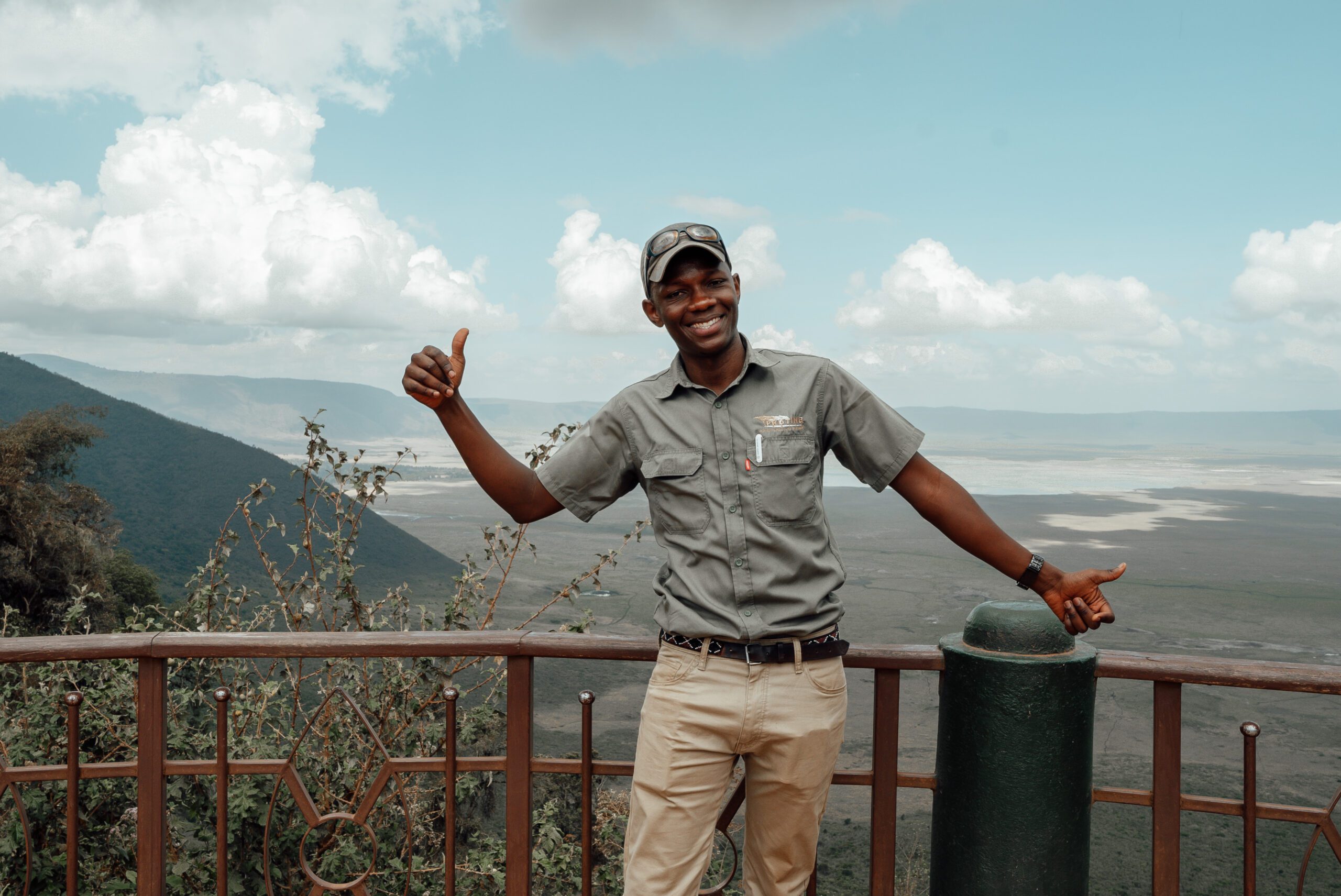 A person in a cap and casual clothing stands by a railing, smiling and giving a thumbs-up, with a vast landscape and cloudy sky in the background—a perfect moment to capture before you book your trip.