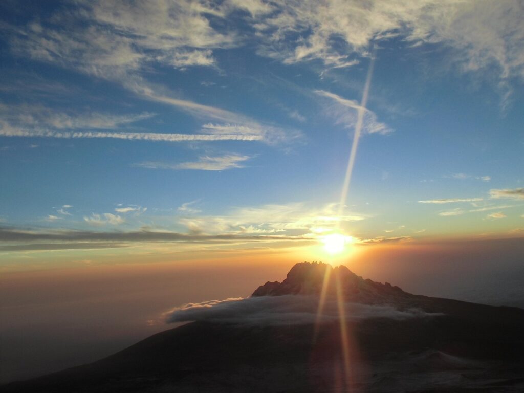 Sunrise over a mountain peak in Tanzania with scattered clouds and a clear sky, emitting rays of light, invites you to visit this breathtaking landscape.