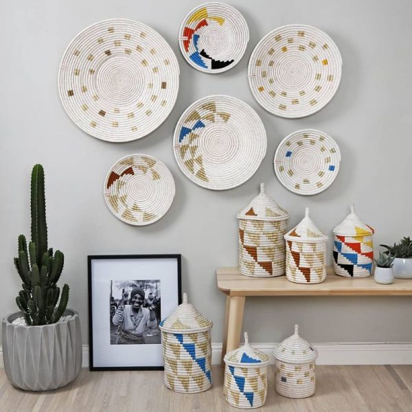 Woven baskets with geometric patterns hang on a wall, embodying a perfect touch for any Holiday Gift Guide. Below, covered baskets and a framed photo sit on a bench, next to a potted cactus.