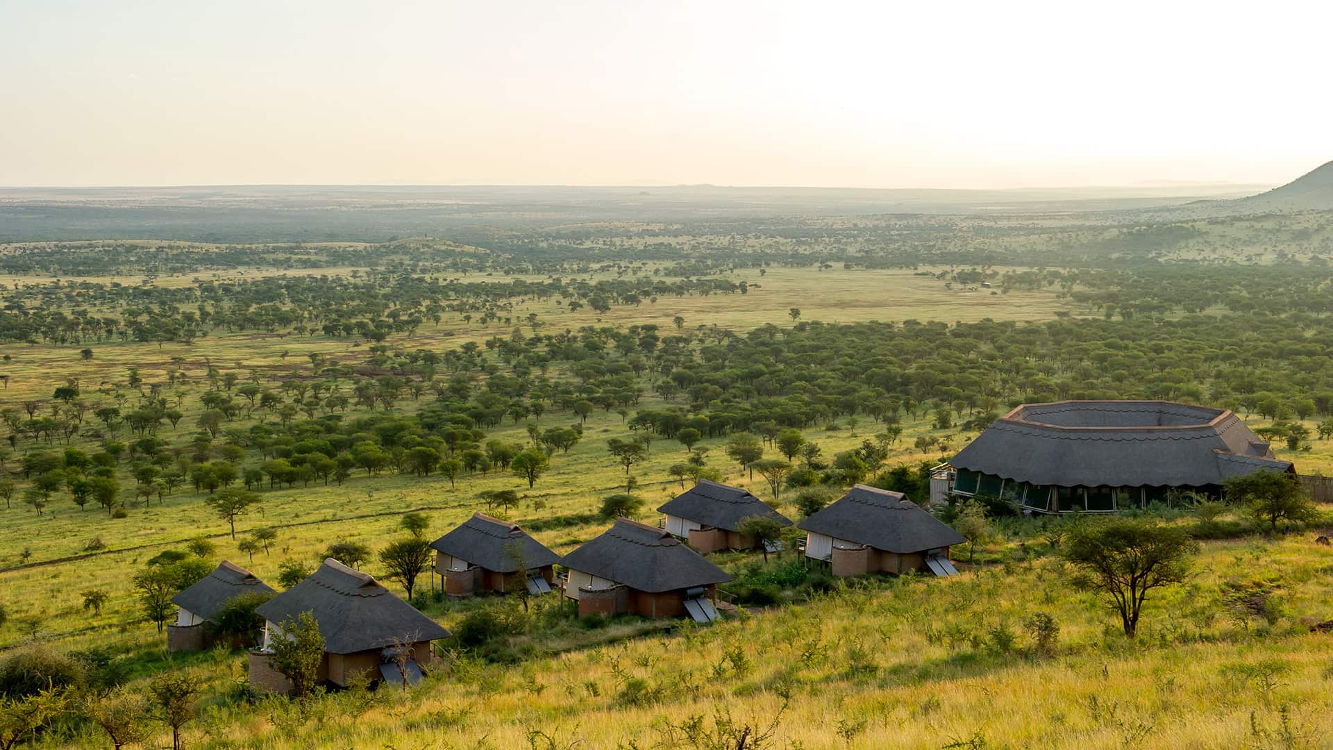 Aerial view of a series of round huts and a larger circular building located in a vast, green savannah landscape.