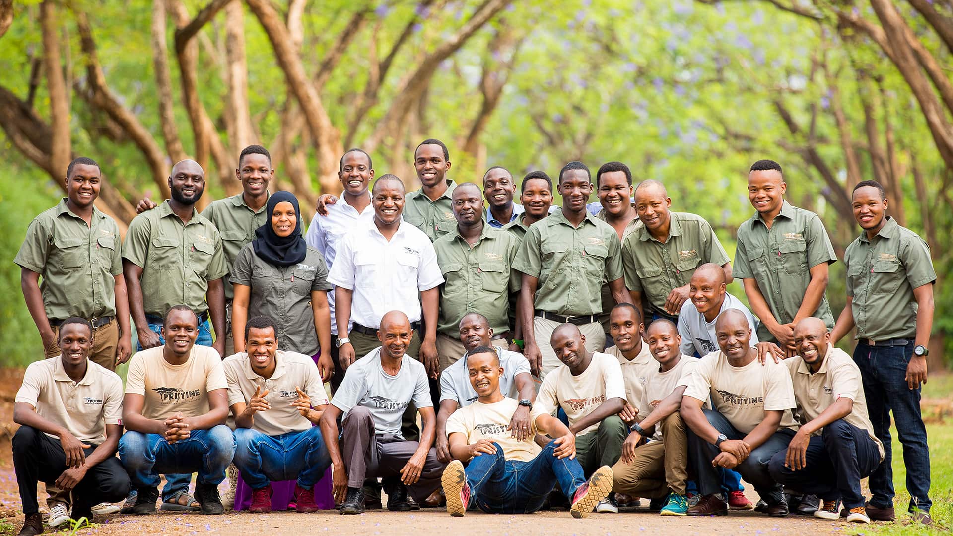 A diverse group of people, wearing uniforms and casual outfits, pose together smiling outdoors with a background of trees.