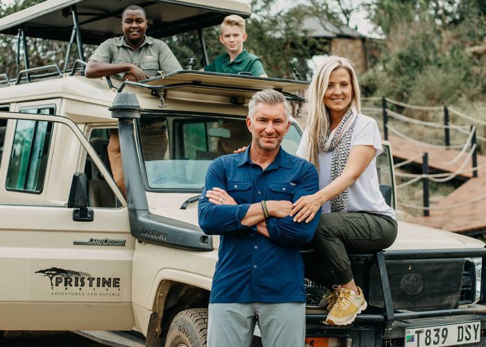 Four people are posed around a pristine safari vehicle; two are standing, and two are sitting. The setting suggests an exciting family adventure in Tanzania, possibly during a 10-day tour through the breathtaking safari parks.