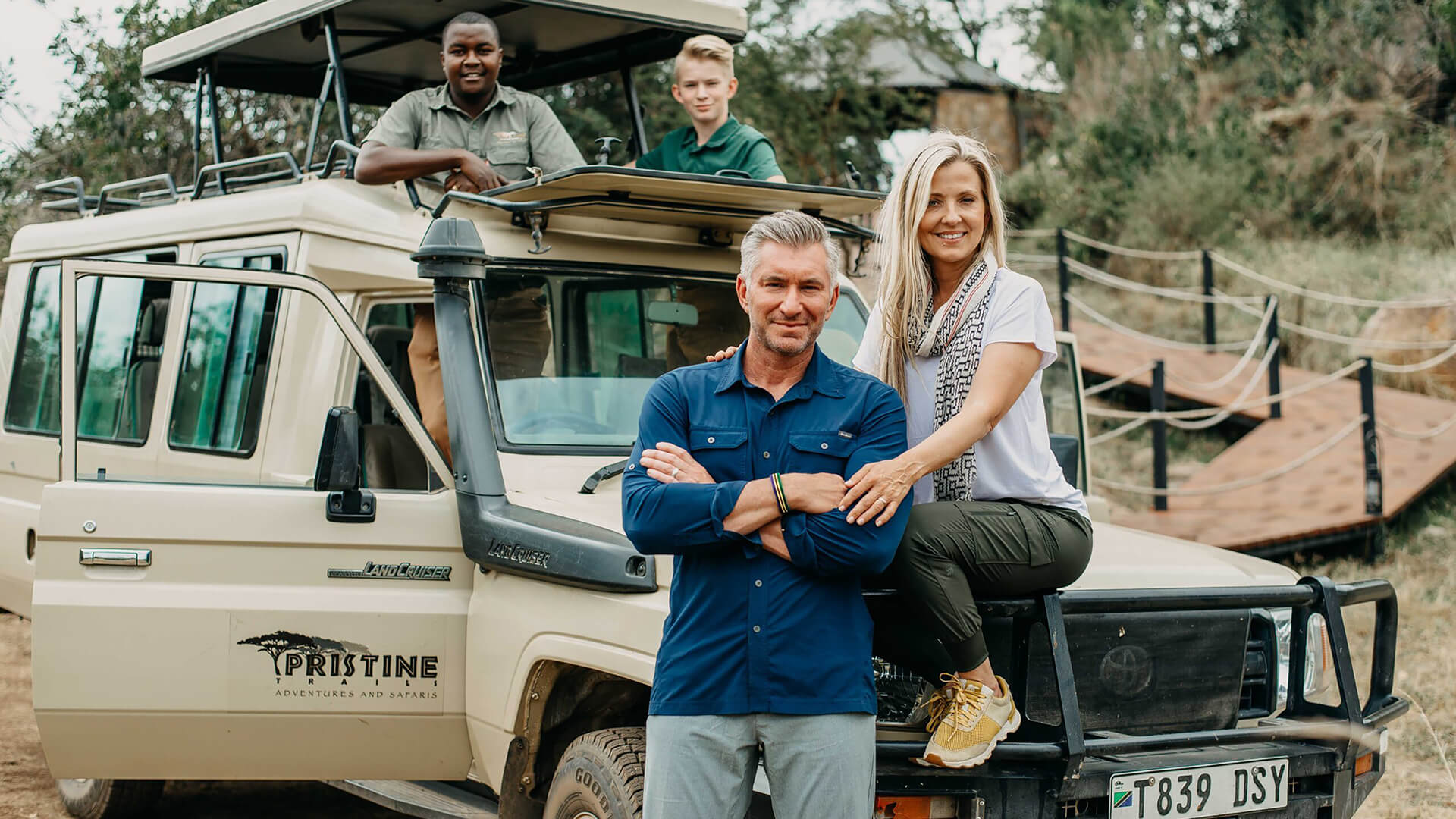 Four people are posed around a pristine safari vehicle; two are standing, and two are sitting. The setting suggests an exciting family adventure in Tanzania, possibly during a 10-day tour through the breathtaking safari parks.