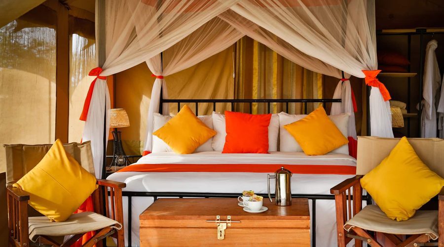 Bed with orange and yellow pillows under a mosquito net canopy, flanked by chairs with matching cushions. A wooden chest and coffee setup are in the foreground.