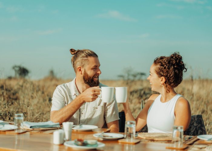 Two people sit at an outdoor table in a field, holding white mugs and smiling at each other, their laughter as warm as the Tanzanian sun. The table is set with plates and glasses, capturing the essence of this perfect stop on their travel itinerary.