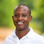 A smiling man, clad in a crisp white shirt, stands against a backdrop of blurred greenery, embodying the spirit of Pristine Adventures and Safaris.