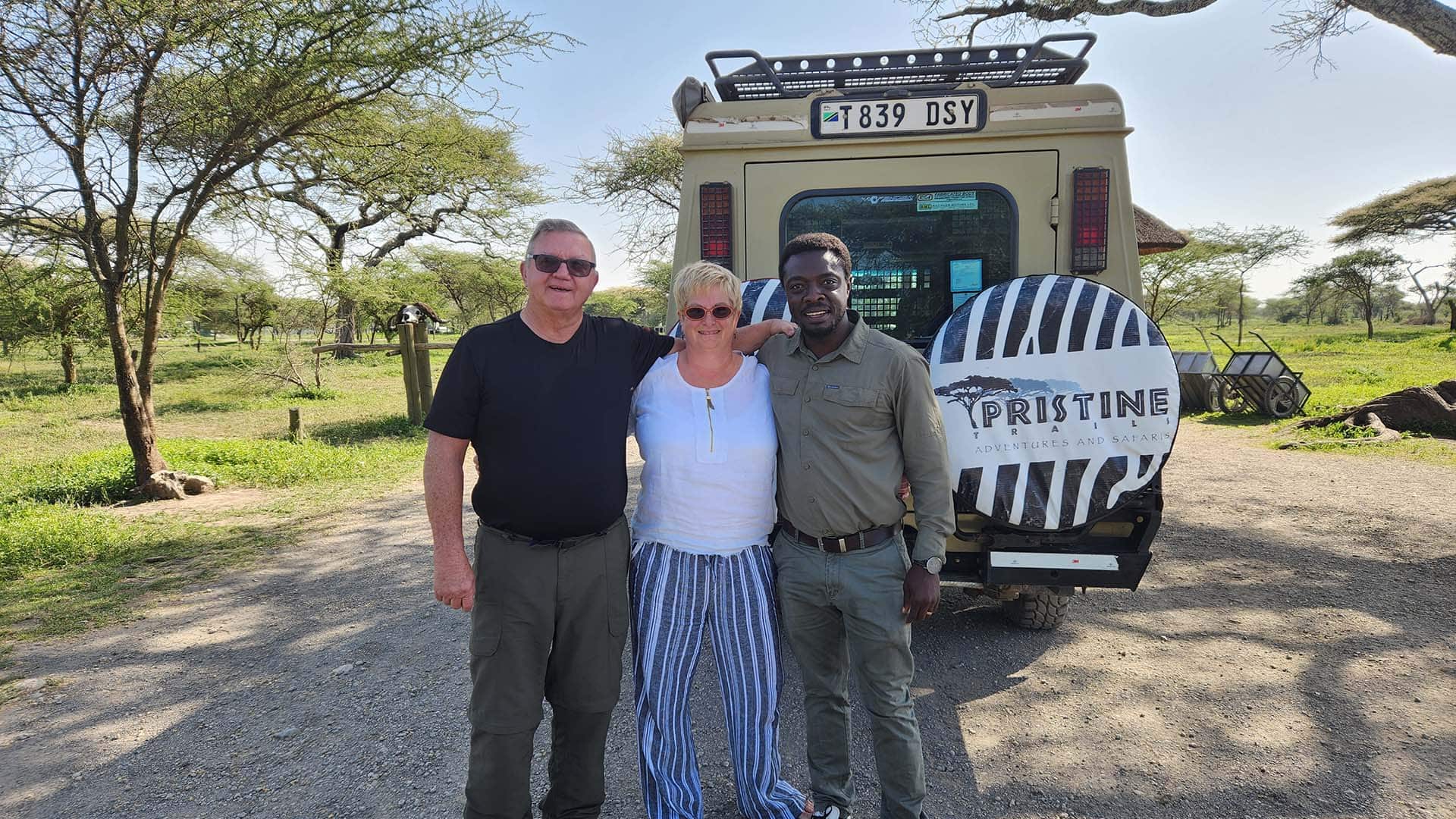 Three people stand before a safari vehicle with a "Pristine" cover in a sunny, grassy landscape dotted with trees, evoking the adventure of luxury lodges nearby.