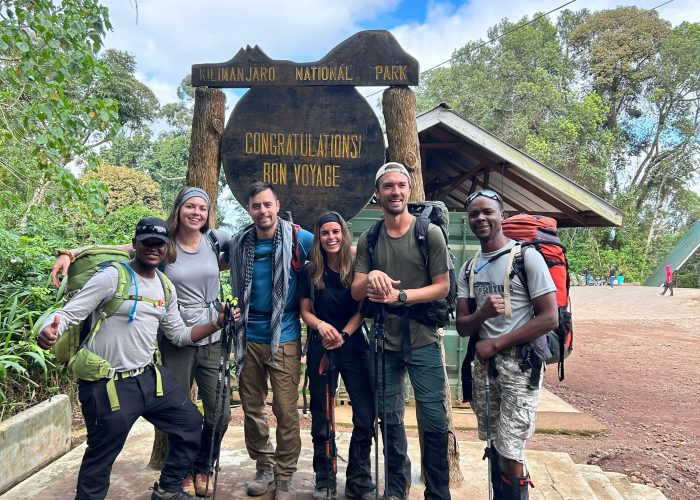 A group of hikers poses in front of the Kilimanjaro National Park sign, holding trekking poles and wearing outdoor gear.
