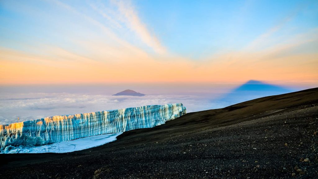 Ice cliff on a mountain slope with a view of clouds and two peaks in the background under a colorful sky at sunset.