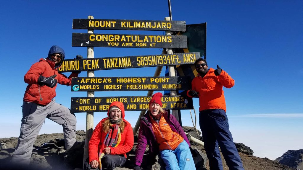Four people in hiking gear stand in front of the Mount Kilimanjaro summit sign, celebrating their achievement on a clear day.