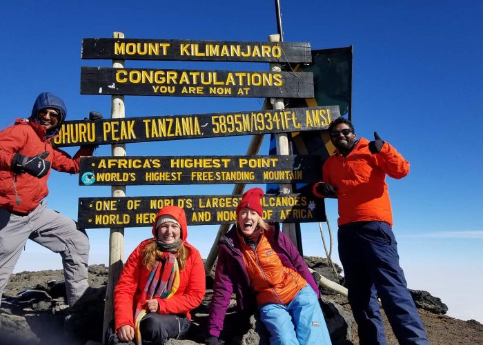 Four people in hiking gear stand in front of the Mount Kilimanjaro summit sign, celebrating their achievement on a clear day.