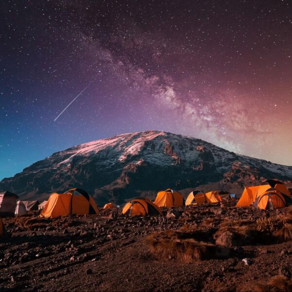 Orange tents set up on rocky terrain under a starry night sky with the Milky Way and a snowy mountain in the background.