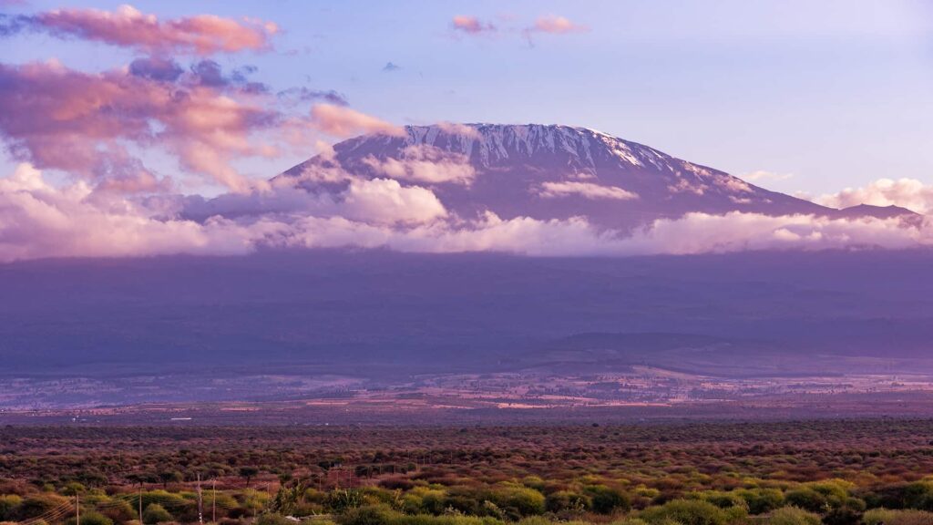 A distant view of a snow-capped mountain peak surrounded by clouds, overlooking a vast landscape of green vegetation and open plains.