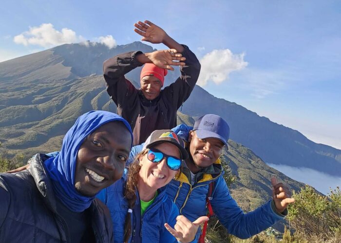 Four people smiling and posing for a selfie atop Mt Meru, capturing the breathtaking landscape during their trekking tour.