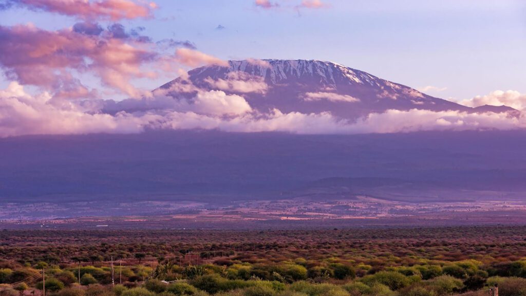 A snow-capped mountain peak rises above a layer of clouds, with a vast plain and sparse vegetation in the foreground—an inspiring scene for first-timers beginning their Kilimanjaro training under a partly cloudy sky.