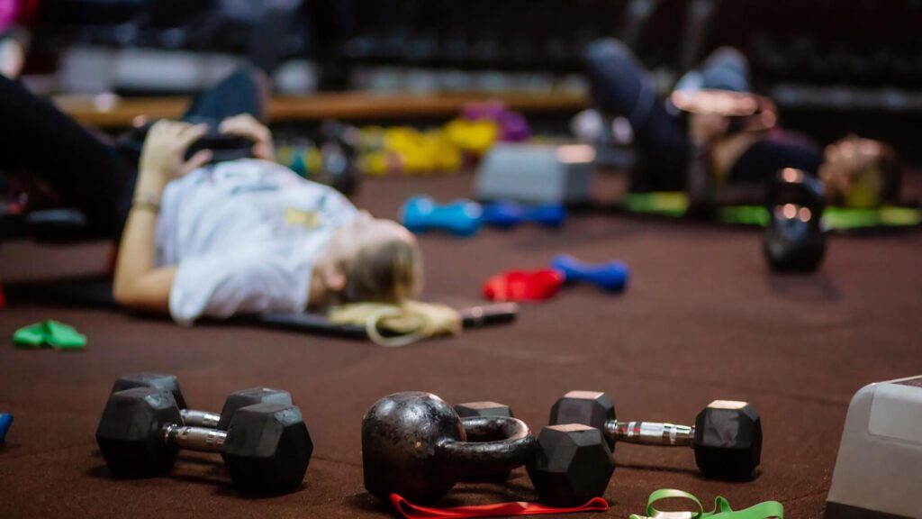 First-timers lying on exercise mats in a gym, with dumbbells, a kettlebell, and resistance bands scattered on the floor—perfect for starting your fitness plan or Kilimanjaro training.