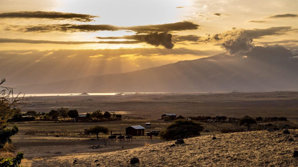 Sun rays break through clouds over a rural landscape with scattered trees, cattle, and small houses—perfect scenery for a first-time safari—with mountains and a body of water in the background.