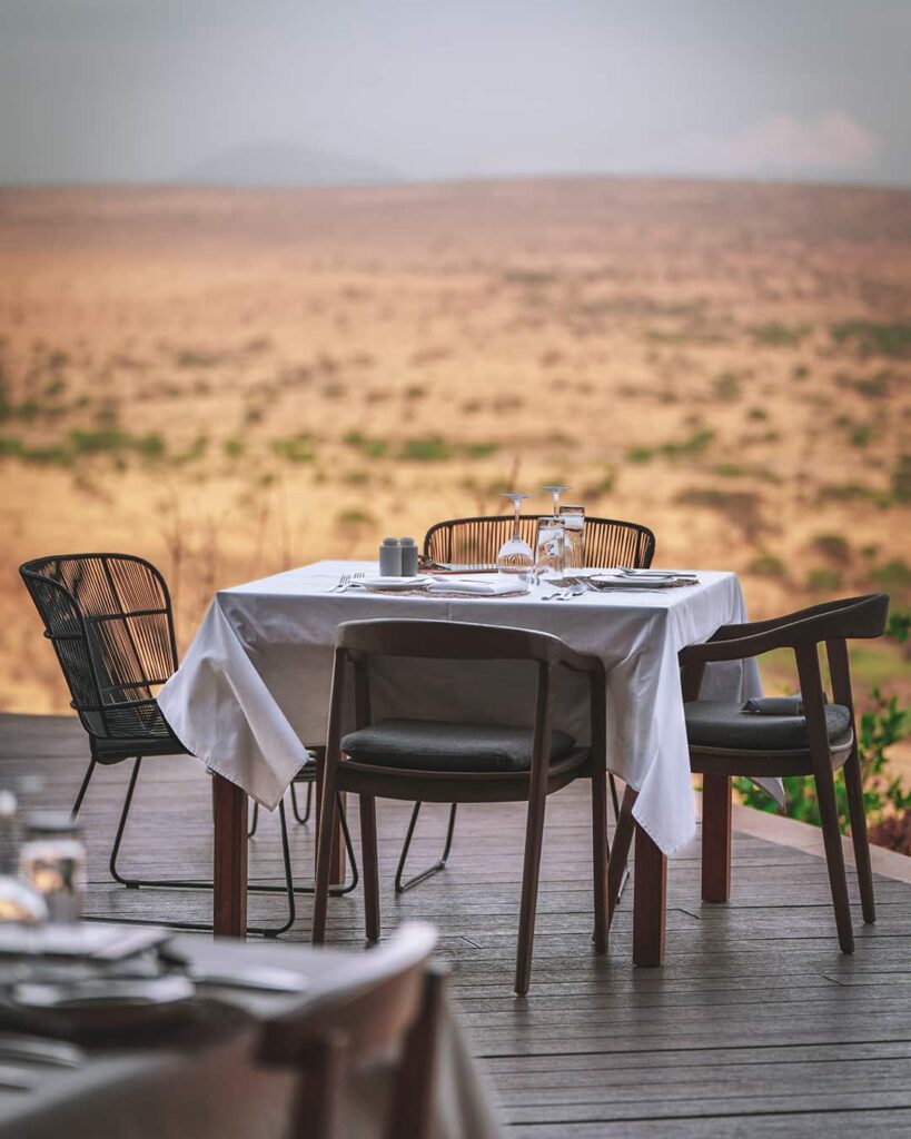 A set dining table with four chairs is arranged on a wooden deck, overlooking a vast, arid landscape under a cloudy sky.