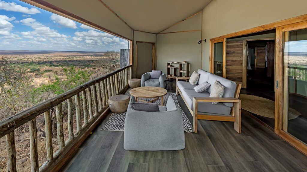 A covered wooden deck with modern outdoor furniture overlooks a vast savanna landscape under a partly cloudy sky. Sliding glass doors lead to an interior room.
