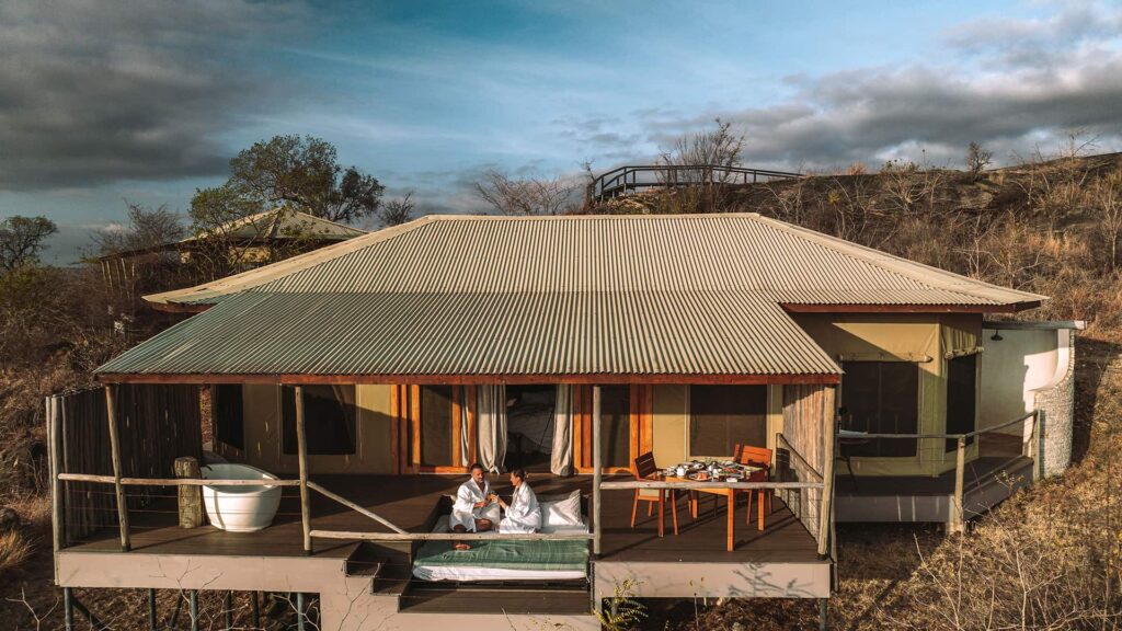 Aerial view of two people in robes sitting on the deck of a luxury cabin with an outdoor bathtub, surrounded by dry vegetation and under a partly cloudy sky.