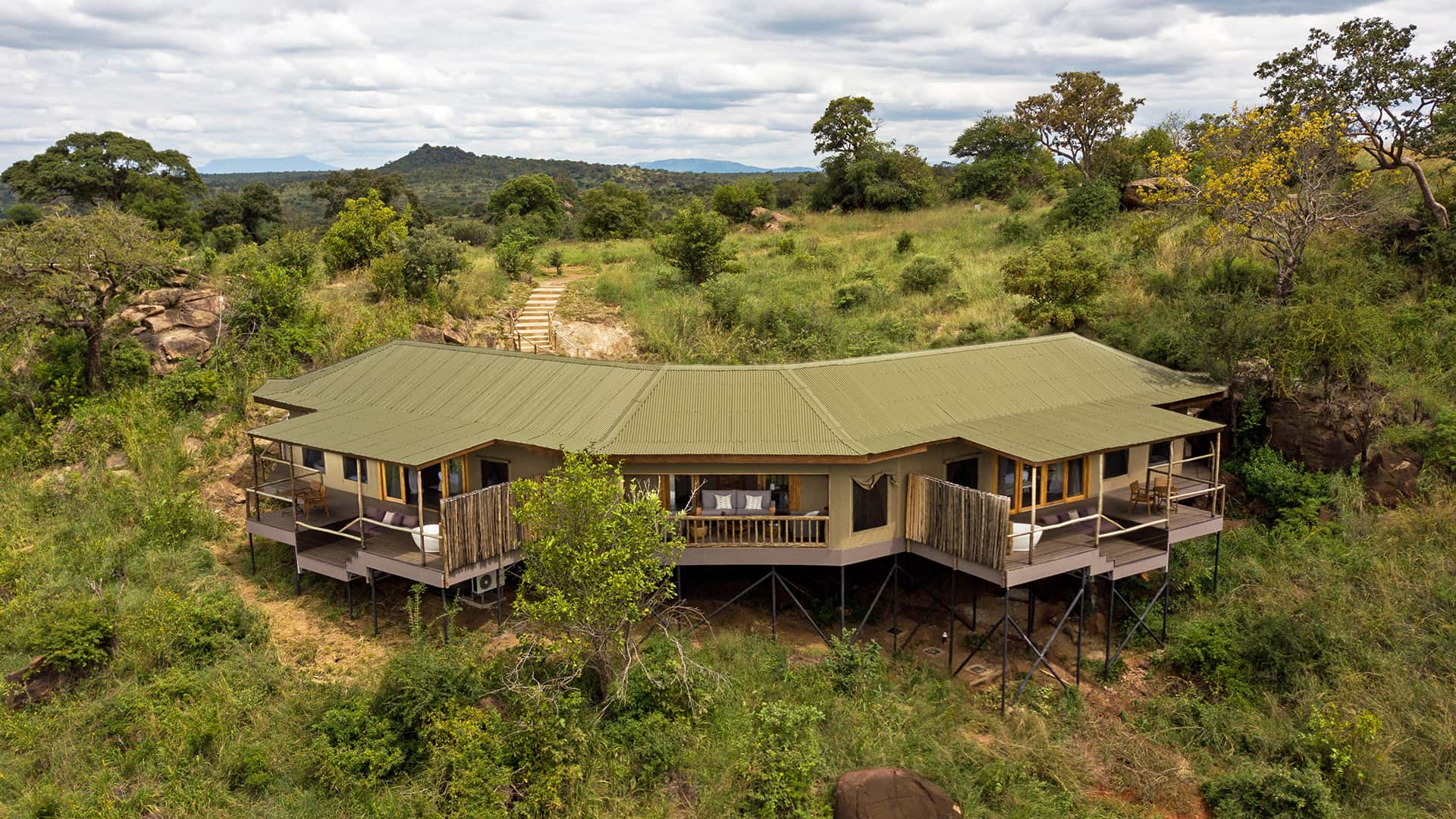 A modern safari lodge with a green roof and wraparound decks is elevated on stilts in a grassy, wooded landscape under a cloudy sky.