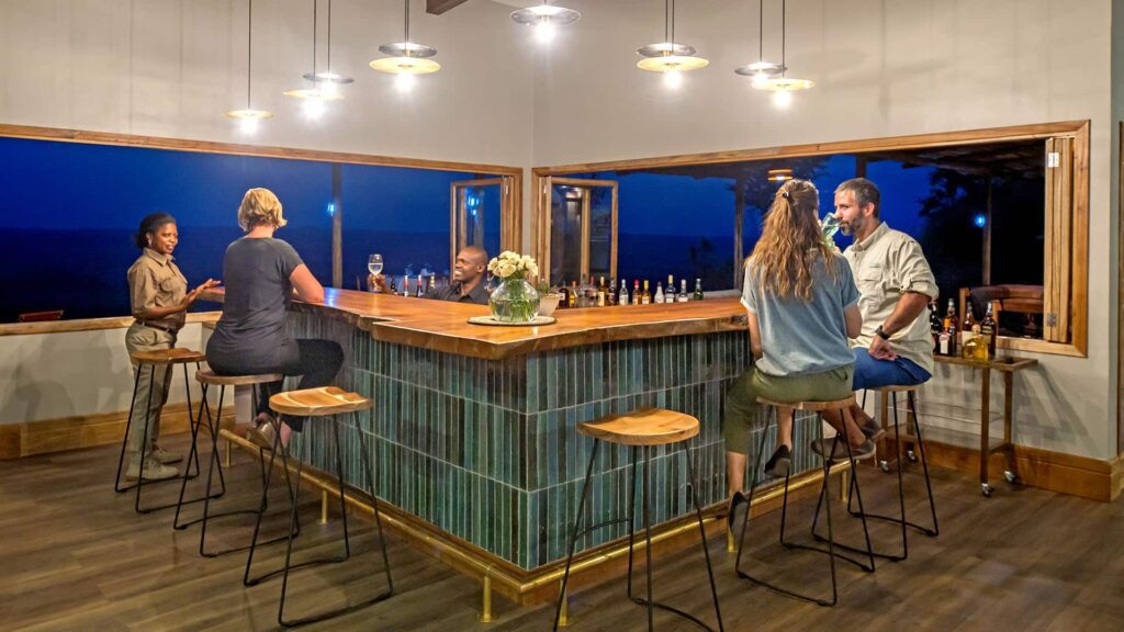 Five people sit and converse around a modern, U-shaped bar in a well-lit indoor space with large windows overlooking a nighttime view.