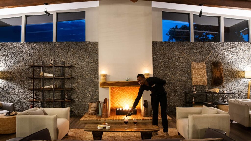 A person places a glass of wine on a coffee table in a modern living room with a lit fireplace, neutral furniture, and stone accent walls at dusk.