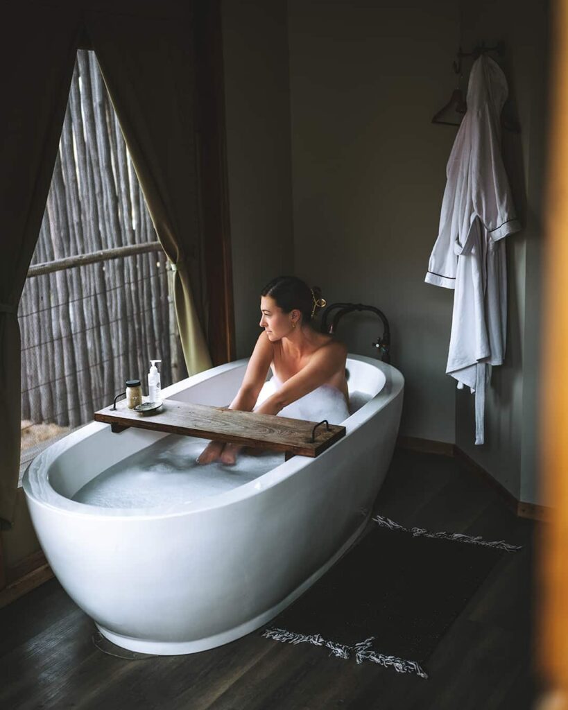 A person sits in a white freestanding bathtub filled with water, looking out a window. A wooden bath tray holds items, and a white robe hangs on the wall nearby.