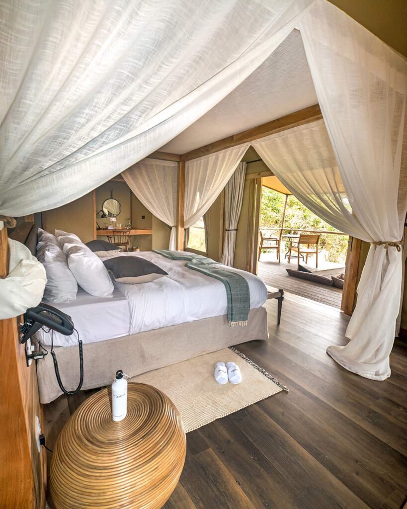 A modern canopy bed with white linens and throw pillows is in a bright room with wooden floors, sheer curtains, a round wooden table, slippers, and an open view to a furnished balcony.