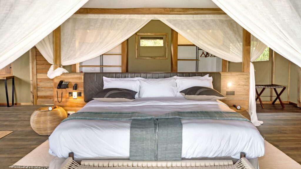 A neatly made bed with white linens and gray accents sits under a canopy in a modern, wooden, open-air room with natural light.
