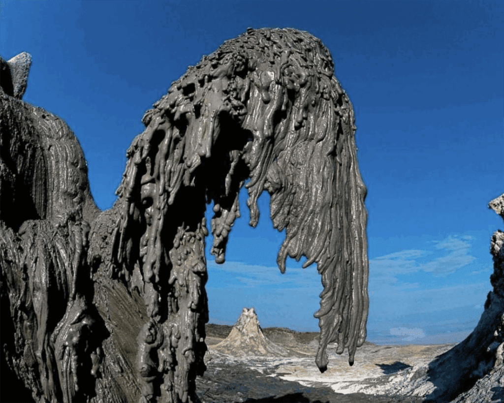 Thick, gray mud flows and hardens into unusual shapes at a mud volcano site in Tanzania under a clear blue sky, with a distant rocky outcrop—an incredible spot to visit for adventurous travel enthusiasts.
