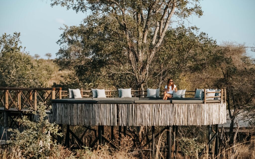 A woman sits on an elevated wooden deck with cushions at Nimali Tarangire, surrounded by trees and overlooking a natural landscape—the perfect setting for a tranquil Tanzania luxury lodge experience.