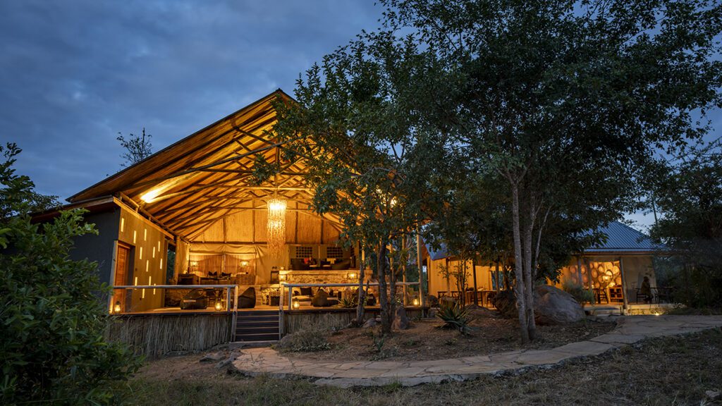 A Tanzania luxury lodge with an open, thatched roof is warmly lit at dusk, surrounded by trees and a stone pathway leading to the entrance—welcome to Nimali Tarangire, your gateway to an unforgettable Tarangire safari.