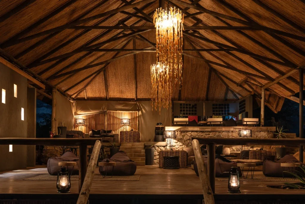 Open-air lodge at Nimali Tarangire with a thatched roof, wooden beams, wicker furniture, hanging gold chandelier, and lanterns glowing on a wooden deck at night.