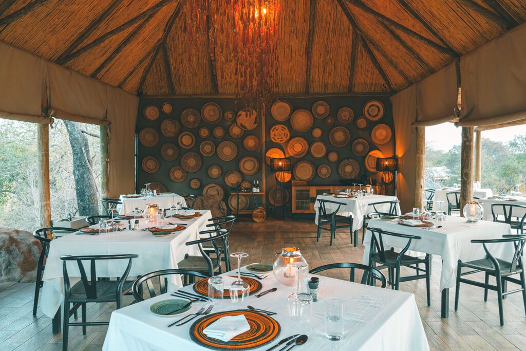 A rustic restaurant dining area at Nimali Tarangire features tables covered in white cloths, black chairs, woven baskets on the back wall, and natural light streaming through large open windows.