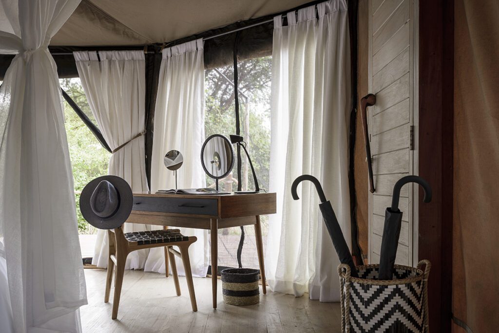 A wooden desk with a mirror, hat, and chair is placed near large windows with sheer curtains; two umbrellas are in a patterned basket on the floor, evoking the tranquil style of Sanctuary Swala in Tarangire National Park.