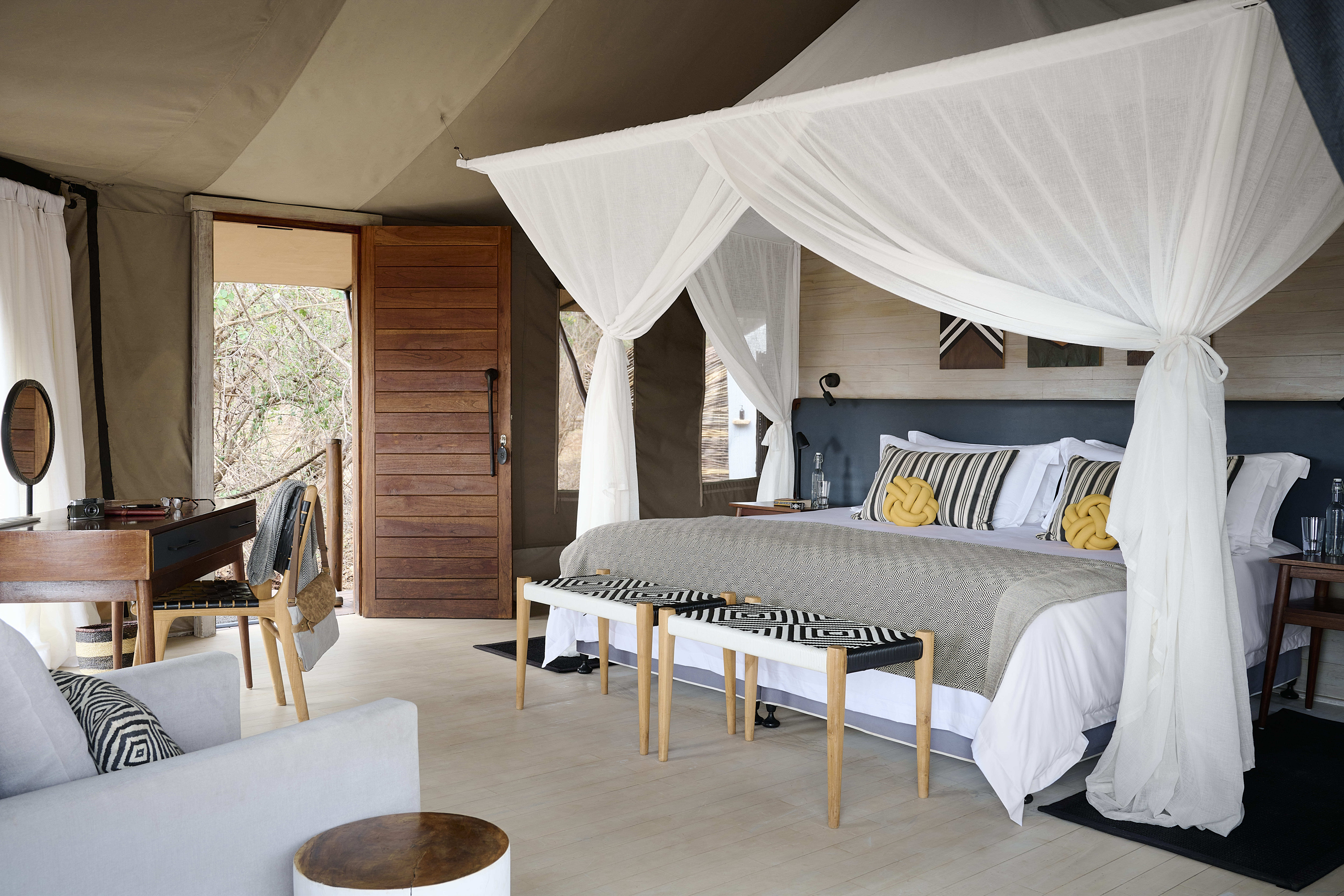 A well-lit bedroom at Sanctuary Swala boasts a canopy bed with neutral bedding, two benches, a desk, a sofa, and wooden decor elements. The open door reveals lush greenery outside, evoking the ambiance of a luxury camp on a Tanzania safari.