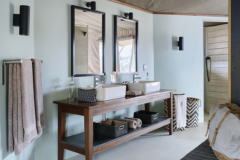 Modern bathroom sanctuary with double sinks, two mirrors, wall-mounted lights, towels, baskets on a wooden vanity, and a Swala laundry hamper on the floor.
