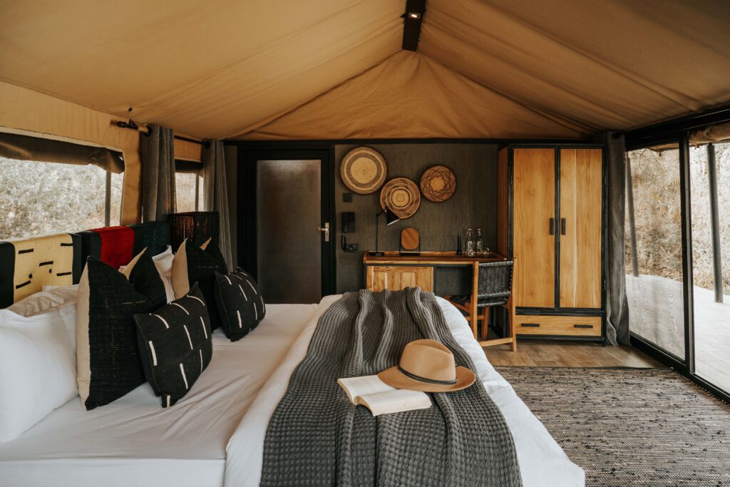 Interior of a safari-style tent at Nimali Tarangire with a neatly made bed, decorative pillows, a hat and book on the blanket, a desk, wardrobe, woven wall decor, and large windows revealing scenic outdoor views.
