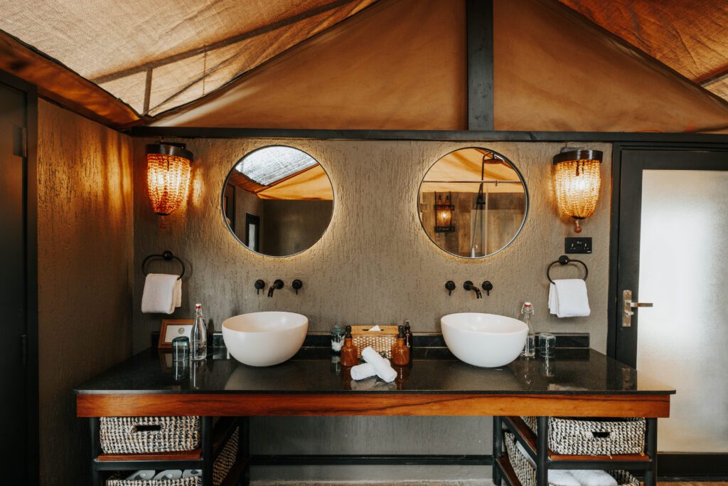A modern bathroom with two round mirrors, two vessel sinks, wall-mounted faucets, and wicker baskets sits atop a black countertop, featuring warm lighting fixtures inspired by the elegant style of Nimali Tarangire.